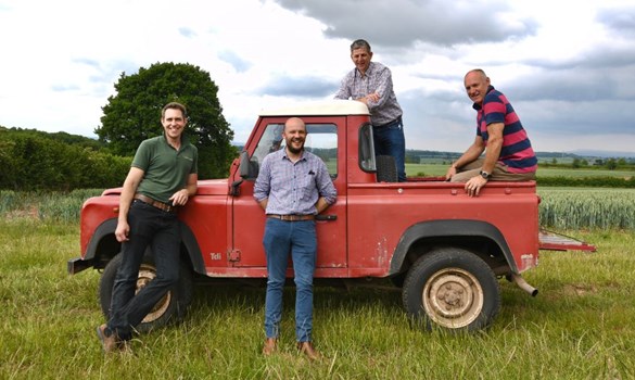 Four men stood on and next to a red land rover in a grassy field